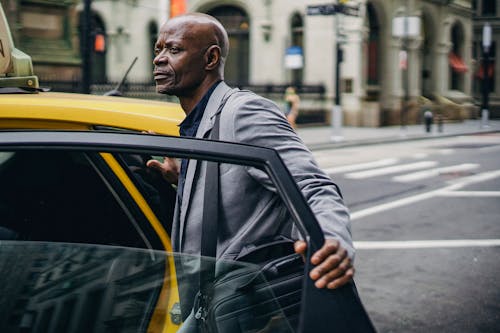 Mature contemplative African American male office worker with briefcase standing on city roadway near cab transport while looking away