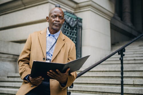 Pensive mature African American male office worker with open folder sitting on staircase handrail while looking away on city street