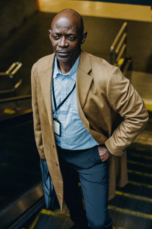 From above of pondering mature African American male office worker standing with hand in pocket on moving stairs and looking away