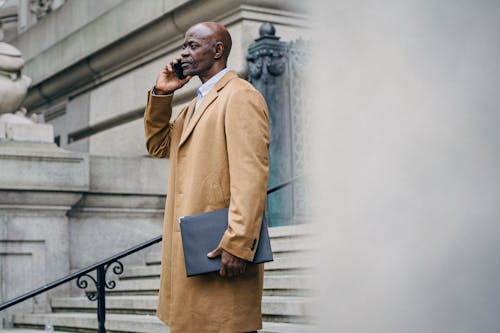 Side view of serious mature African American male entrepreneur with folder speaking on cellphone on urban staircase while looking forward