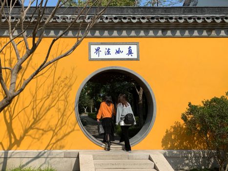 Two women walk through a circular entrance in a vibrant yellow wall in Taizhou Shi, China.