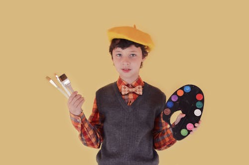 Creative boy showing palette with paints and brushes against yellow background