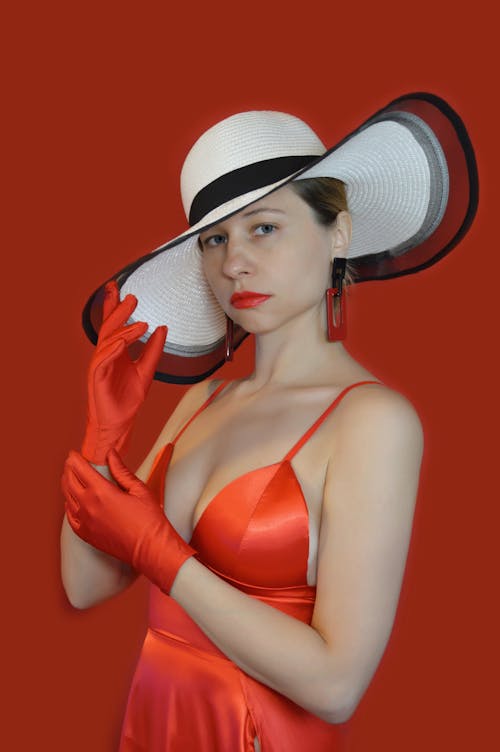 Free Serious female in stylish red dress and hat standing with raised hands and looking at camera against red background Stock Photo