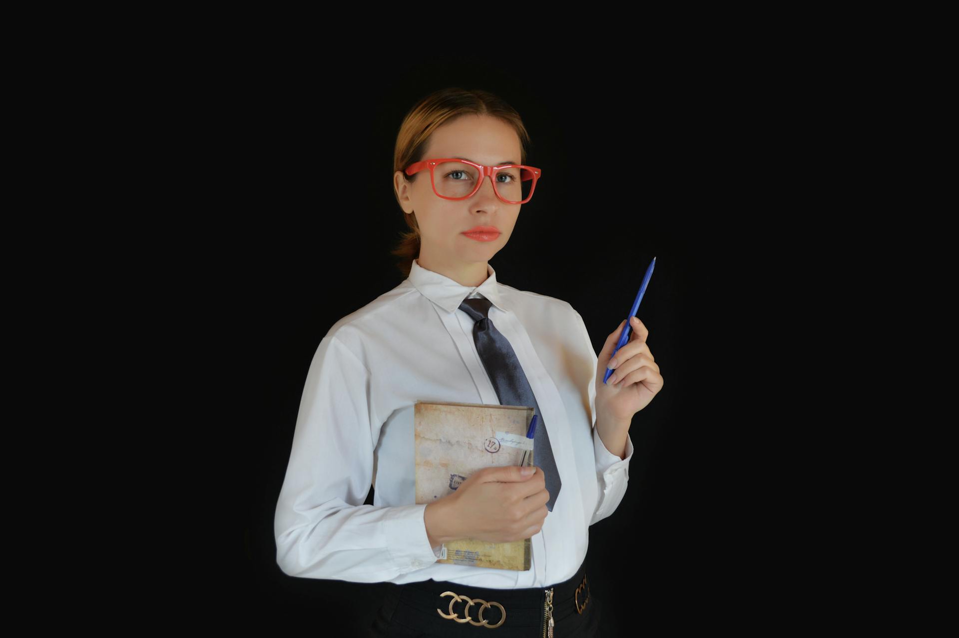 Clever young female executive in formal clothes and eyeglasses standing with diary and pen on black background and looking at camera thoughtfully