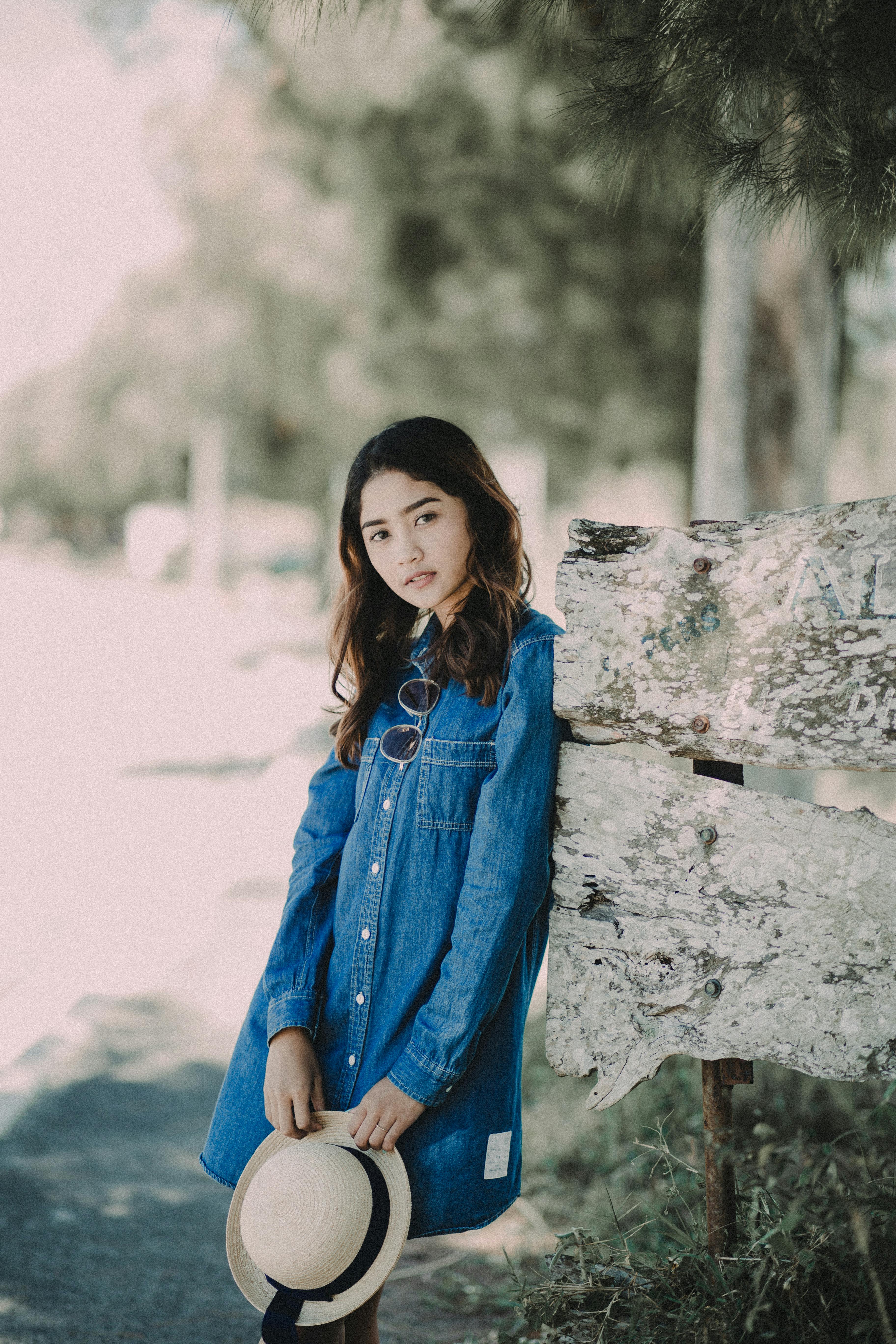 Portrait of a young woman in a denim dress outdoors, holding a hat and leaning against a tree.