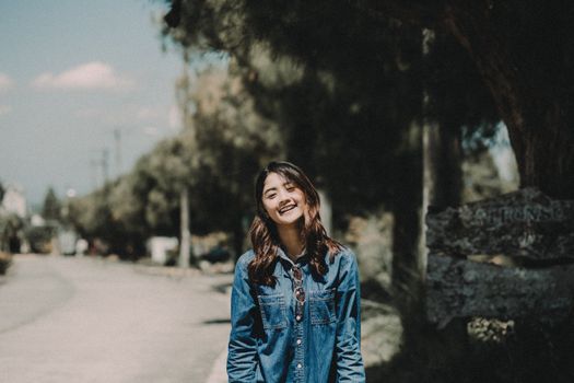 Asian woman smiling in denim jacket standing outside under a tree on a sunny day.