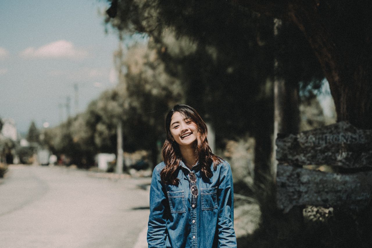 asian woman smiling in denim jacket standing outside under a tree on a