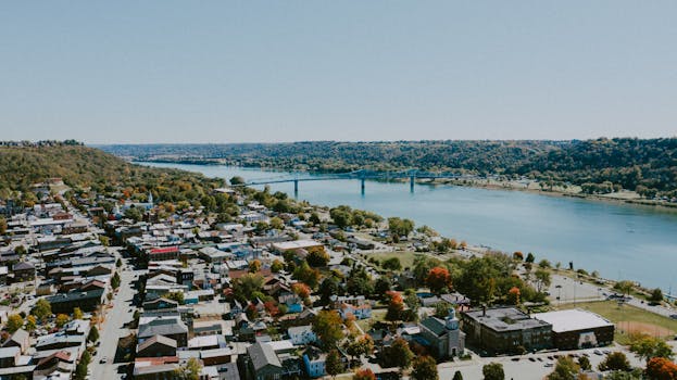 Drone view of small city with houses and buildings near river and green trees growing on hills near bridge under cloudless sky in sunny summer day in countryside