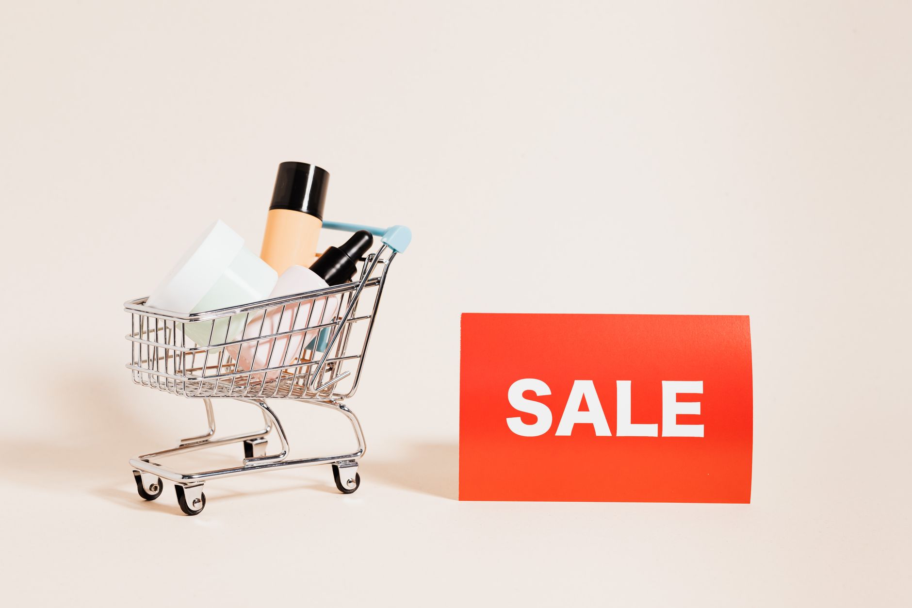 A miniature shopping cart filled with products beside a bright sale sign, ideal for retail and marketing concepts. (Photo by www.kaboompics.com on Pexels)