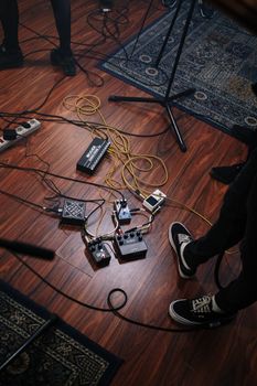 A view of audio equipment and pedals on the wooden floor of a music studio with musicians' legs visible.