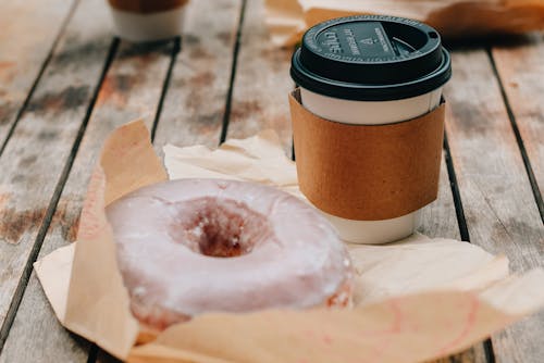 Tasty sweet chocolate donut and takeaway cup of coffee placed on wooden surface in daytime