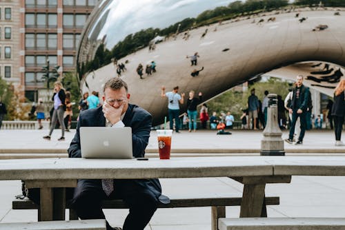 Free Concentrated businessman in suit touching face and sitting at table with laptop and takeaway drink against modern art sculpture Cloud Gate in Chicago in daytime Stock Photo