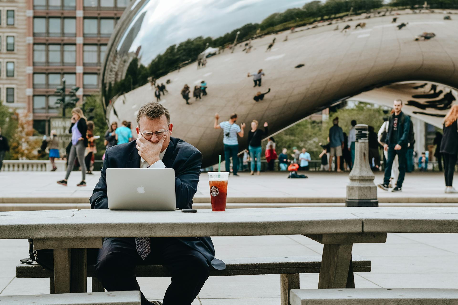 Concentrated businessman in suit touching face and sitting at table with laptop and takeaway drink against modern art sculpture Cloud Gate in Chicago in daytime