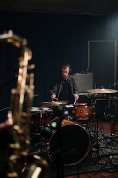 Musician playing drums during rehearsal in a dimly lit studio. A saxophone is in the foreground.