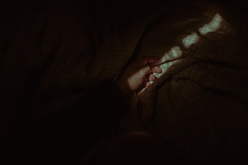Free From above crop anonymous person hand lying on cozy bed with soft blanket in dark bedroom Stock Photo