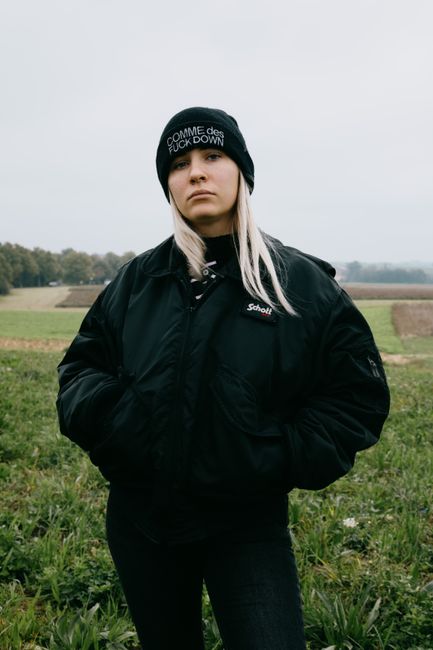 Thoughtful young female in hat and warm coat looking at camera with hands in pockets while standing in meadow under gray cloudless sky