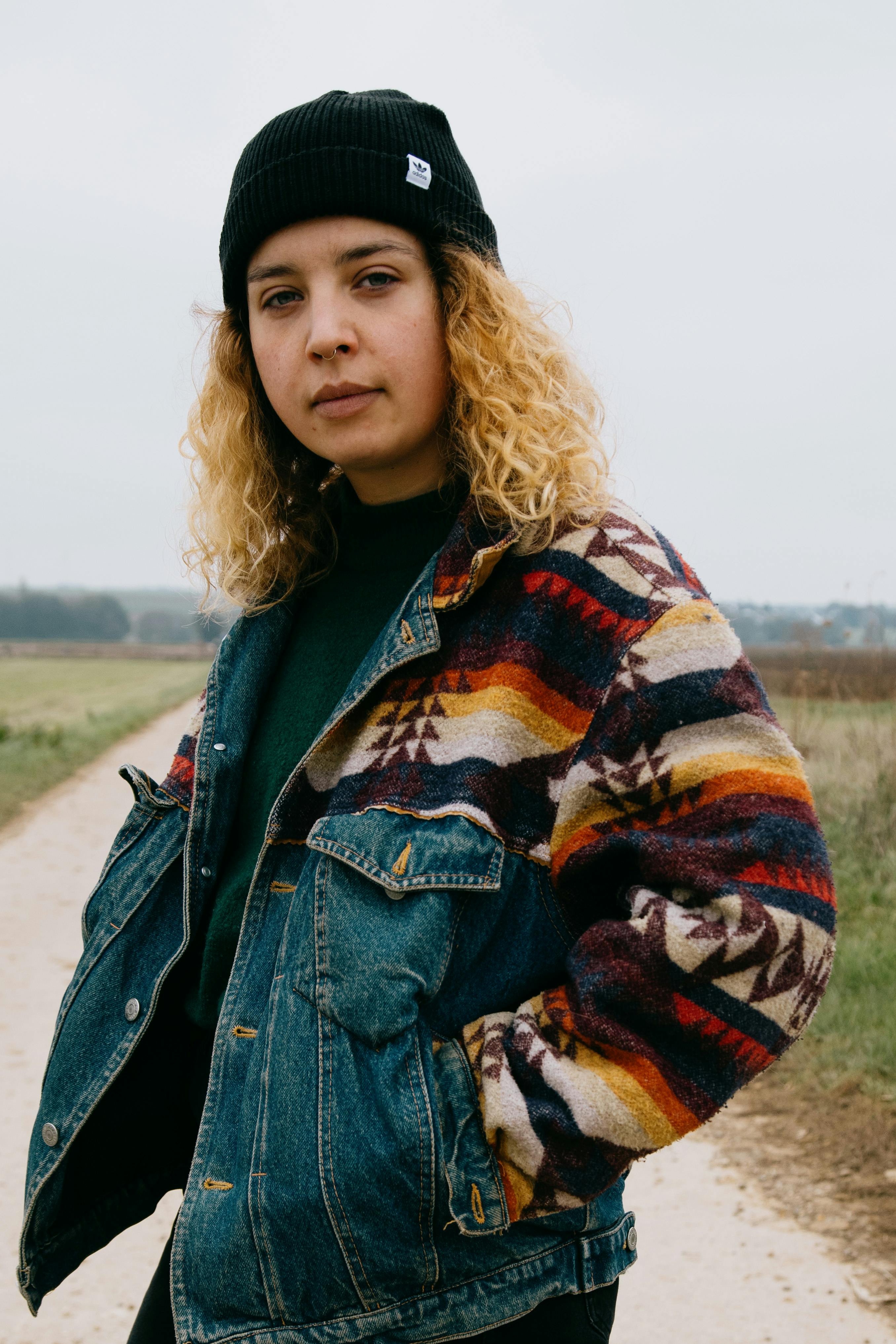 Thoughtful female with curly hair in black hat and bright multi colored denim coat looking at camera with hands in pockets in daylight
