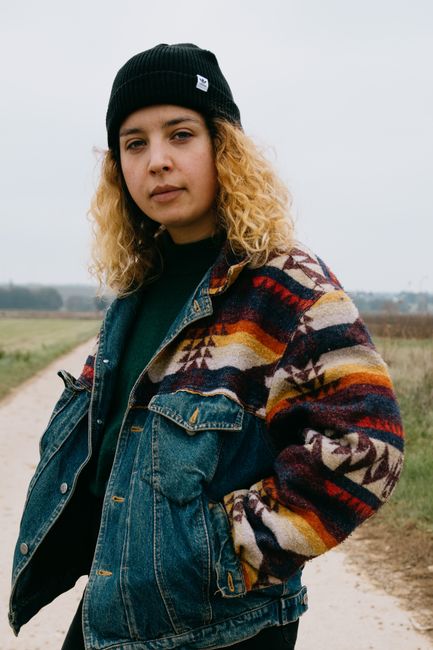 Thoughtful female with curly hair in black hat and bright multi colored denim coat looking at camera with hands in pockets in daylight