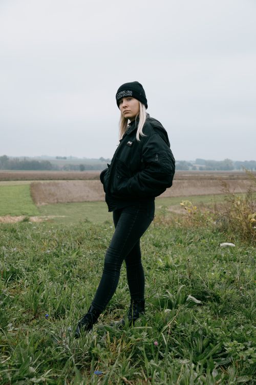 Free Full body of pensive young blond haired female in warm black clothing standing on grass and looking at camera in field under grey sky in autumn Stock Photo