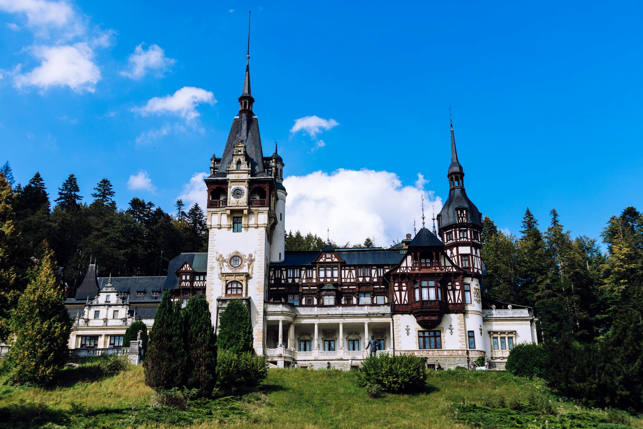 Elegant castle with intricate wood and stone architecture, tall spires, and a clock tower, surrounded by lush greenery under a blue sky.