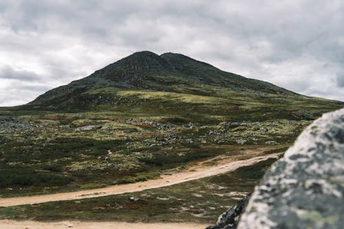 High mountain located in grassy terrain near rural road under cloudy gray sky