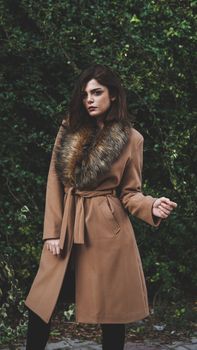 Sophisticated woman in a stylish fur-collared coat standing outdoors against lush greenery.
