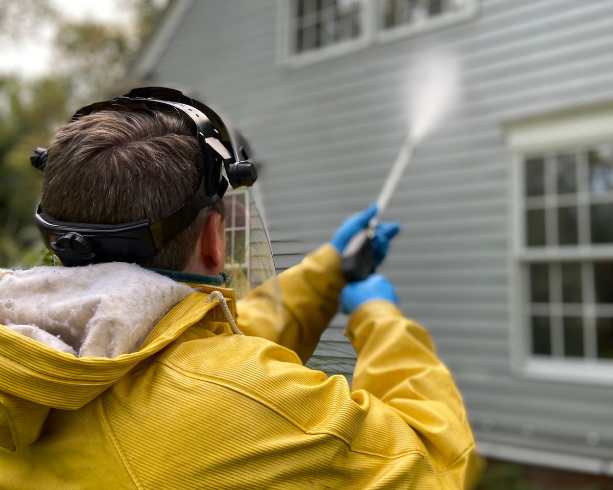 Person cleaning house siding with a pressure washer, wearing protective gear.