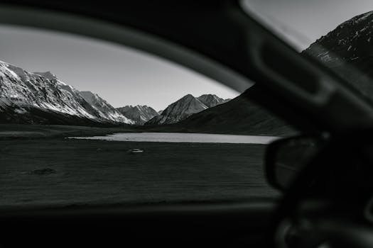 Black and white view of snowy mountains and a calm pond from inside a car.