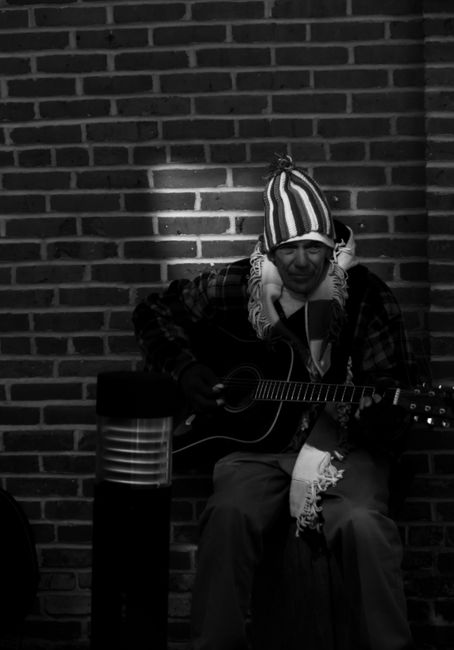 Black and white of man with guitar wearing hat and scarf while sitting near brick wall in street in shade
