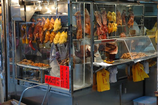 A vibrant street food market stall showing various roasted meats hanging inside a glass cabinet.