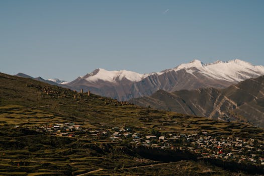 Picturesque village nestled in lush mountains under a clear sky with snowcapped peaks.