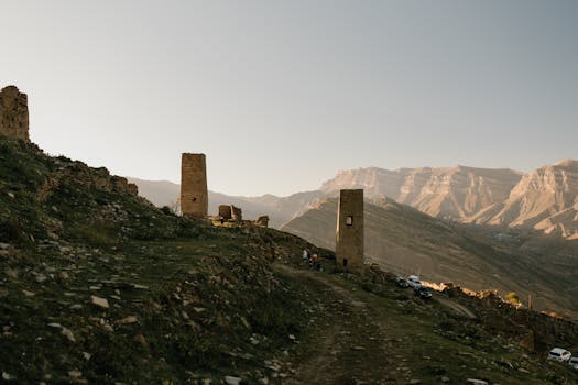 Explore ancient tower ruins in a breathtaking mountain landscape at sunset, with a path leading through history.