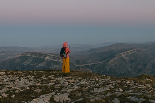 A woman in a hijab and bright pants overlooks a vast mountain landscape at sunset.