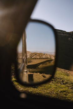Reflection of mountainous landscape in car side mirror under clear skies.