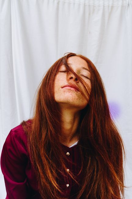 Pensive young redhead female with closed eyes and freckles in casual clothes relaxing against white curtain