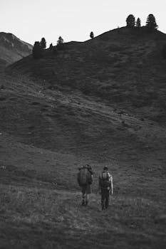 Two hikers traverse a serene mountain path in Cesana Torinese, captured in grayscale.