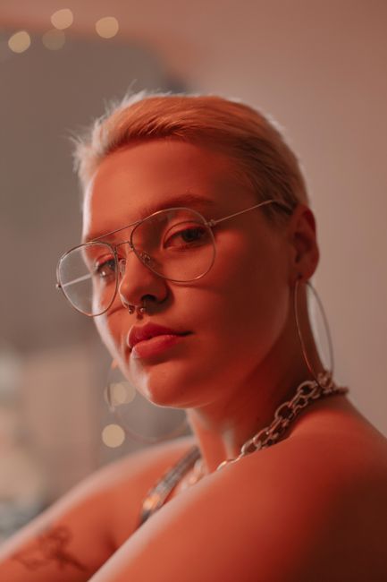 Close-up portrait of a stylish woman with eyeglasses and nose piercing in warm lighting.