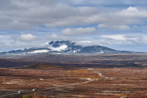 From above of vehicles driving on curvy asphalt roadway among empty terrain leading to high snowy mountain