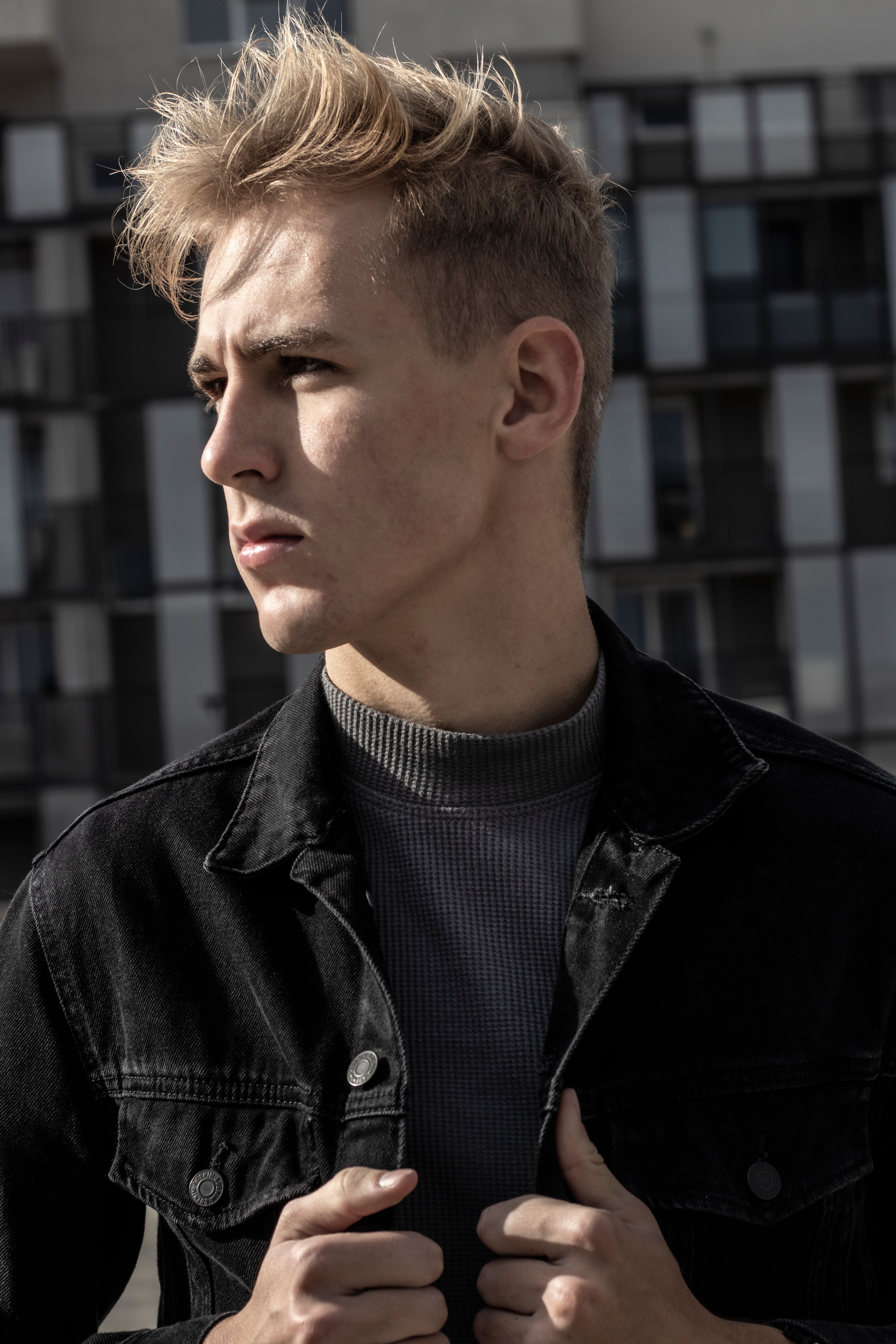 Confident young man in a black jacket posing outdoors with modern architecture backdrop.