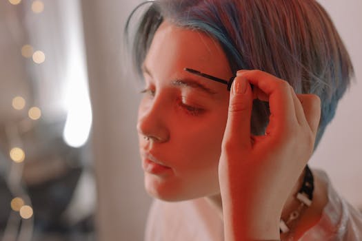 A young woman with dyed hair applying makeup to her eyebrows. Artistic lighting creates a warm ambiance.