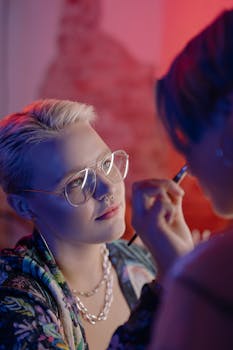 A makeup artist with short hair applies eye makeup indoors. Soft lighting enhances the scene.