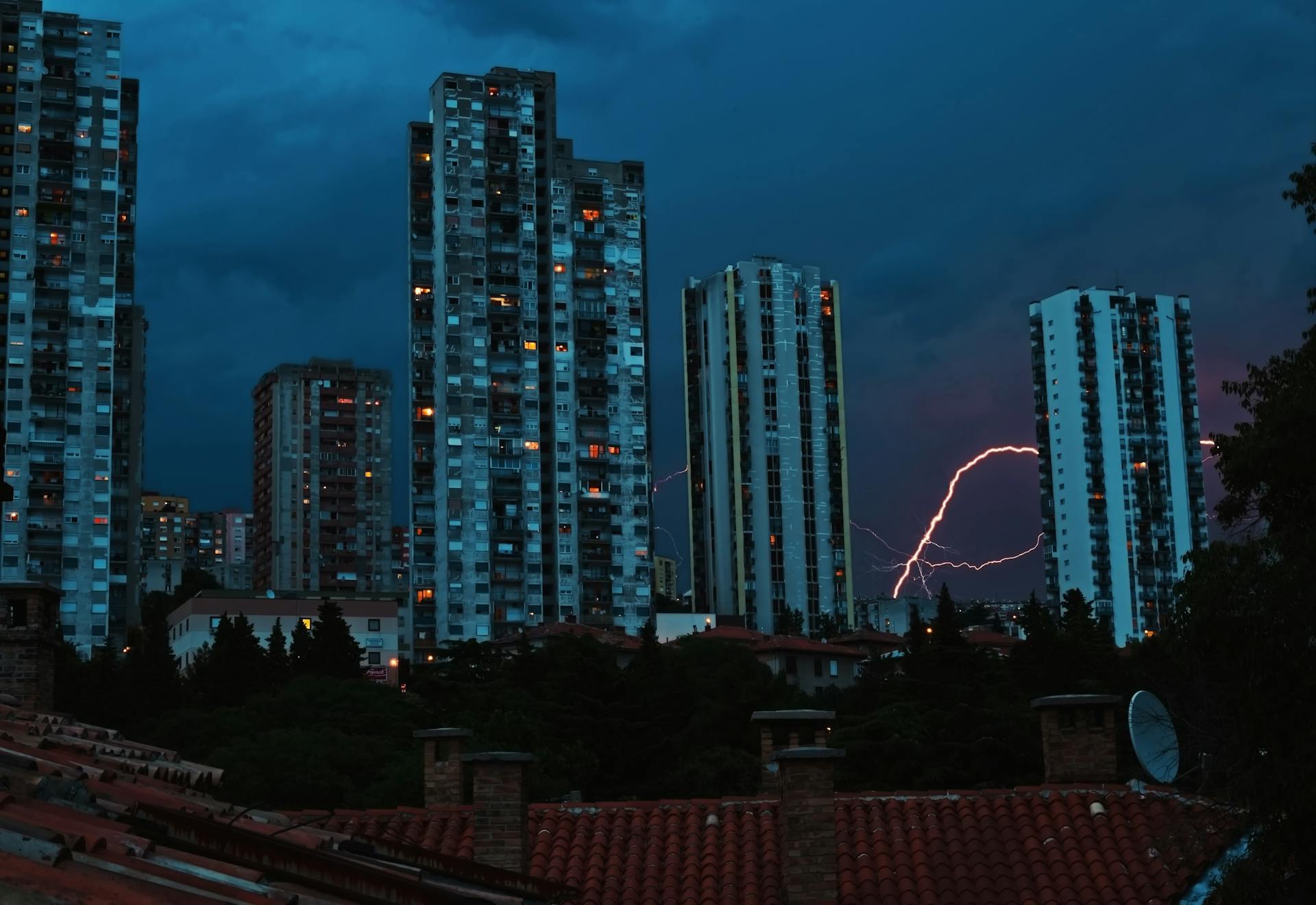 High-rise city buildings illuminated under a dramatic thunderstorm with lightning striking the night sky.