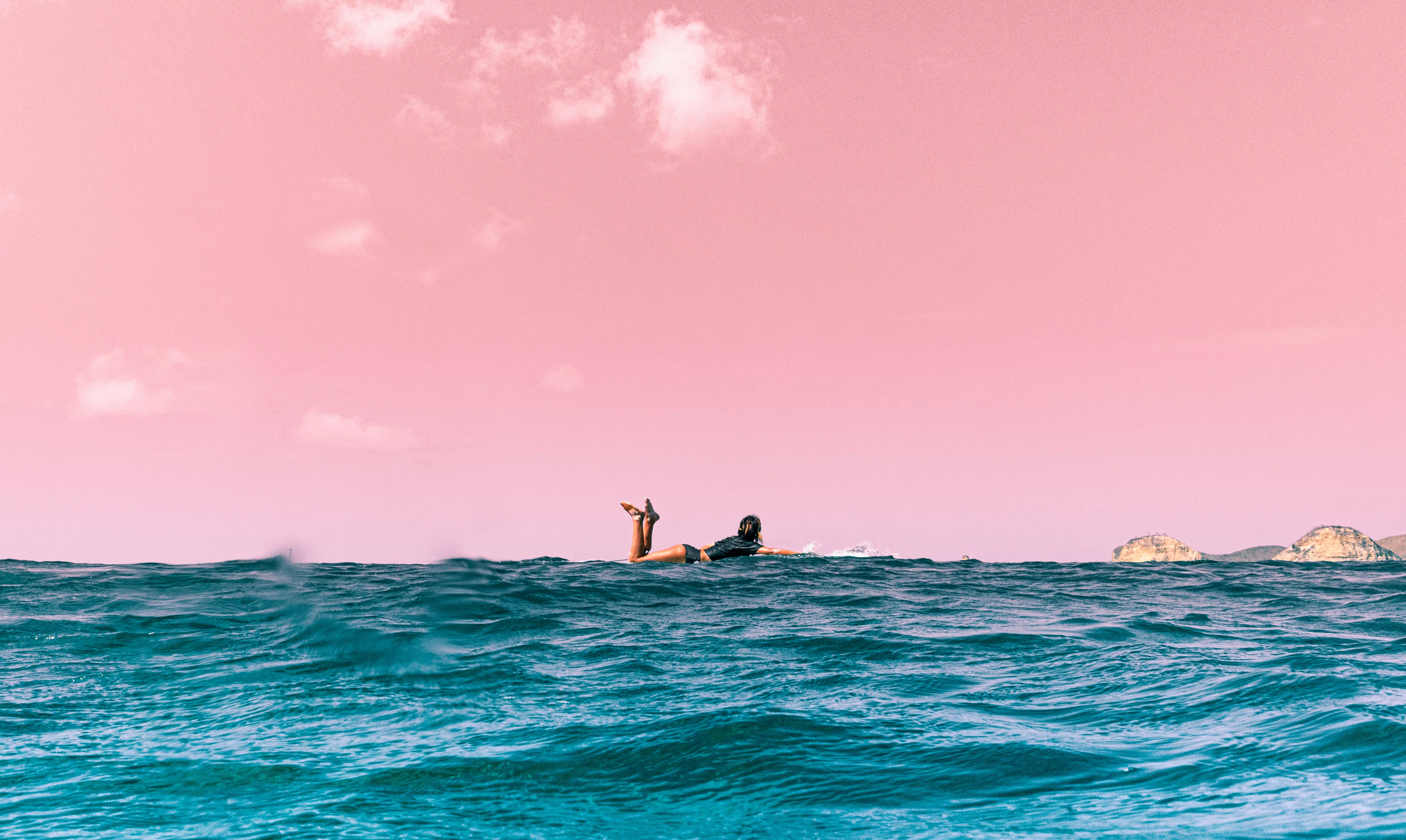 A surfer enjoys the ocean under a striking pink sunset, capturing a tranquil moment on the water.