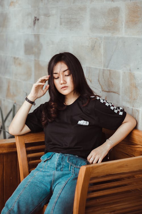 Pensive young Asian female in casual wear sitting on wooden chair outside stone building and touching head