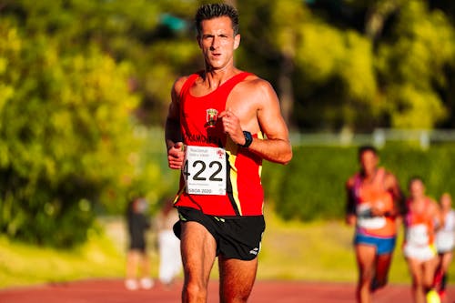 Young focused muscular male athlete in bright sportswear running while participating in athletic contest with unrecognizable rivals and looking at camera