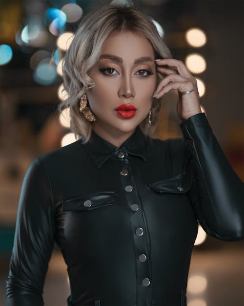 Attractive female with stylish hairstyle and red lips in black leather clothes holding hand on head and looking at camera