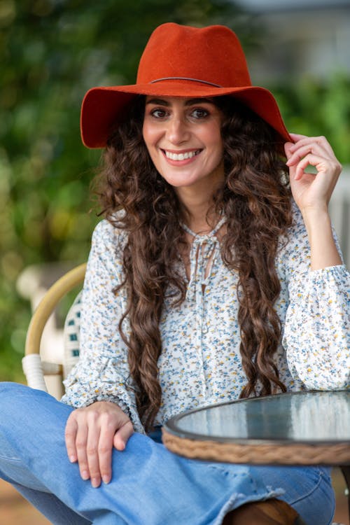 Young happy female in casual clothes touching hat while looking at camera and sitting on chair in street cafeteria during rest