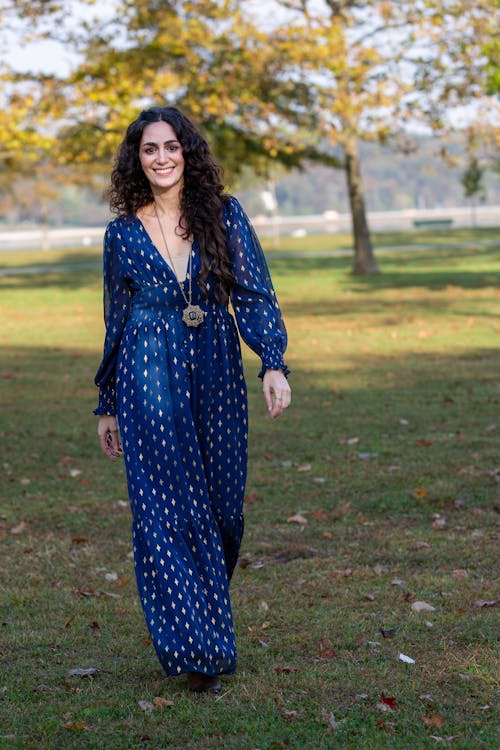 Full length of optimistic female in casual blue dress with toothy smile looking at camera while walking in park during weekend