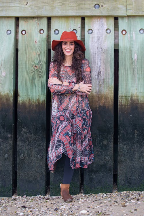 Full body of happy ethnic female with toothy smile and folded hands wearing hat looking at camera