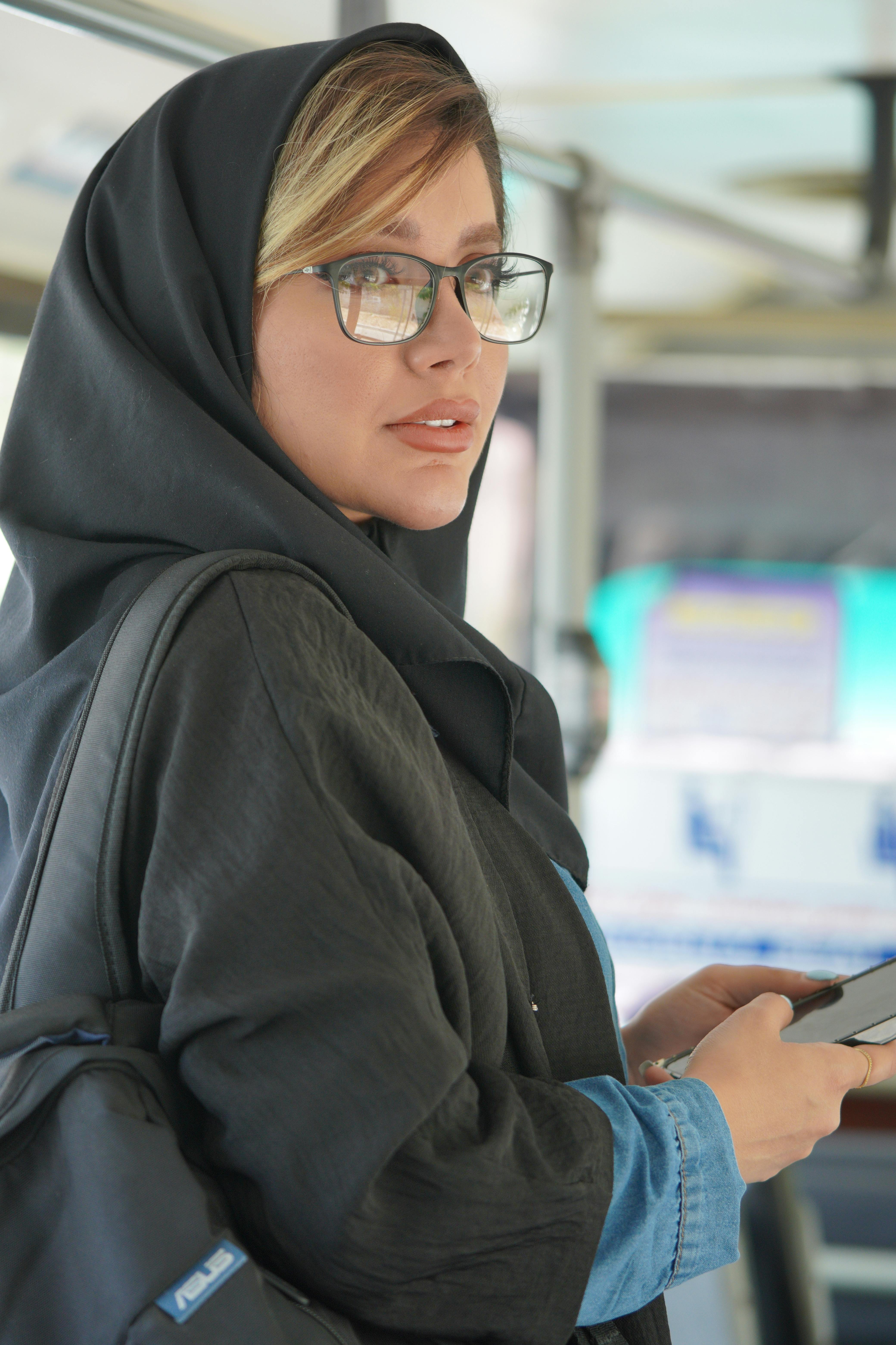 Middle Eastern woman in hijab wearing glasses, using smartphone, inside bus.